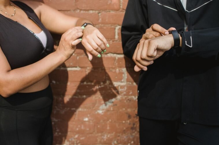 Two people in sportswear checking their fitness trackers outdoors against a brick wall.