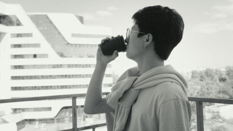Side view of a man sipping coffee on a balcony, overlooking a modern cityscape.