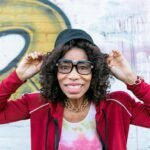 Senior woman smiling playfully wearing stylish glasses and cap in front of graffiti wall.