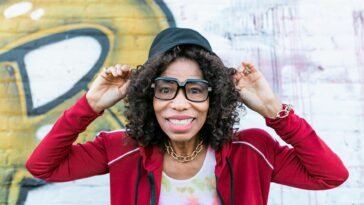 Senior woman smiling playfully wearing stylish glasses and cap in front of graffiti wall.