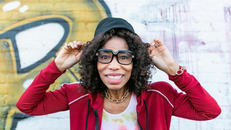 Senior woman smiling playfully wearing stylish glasses and cap in front of graffiti wall.