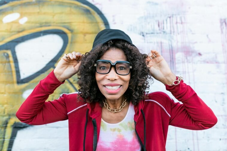 Senior woman smiling playfully wearing stylish glasses and cap in front of graffiti wall.