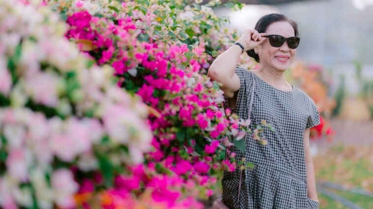 Joyful senior woman wearing sunglasses next to a colorful flower bush outdoors.
