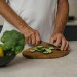 Close-up of adult slicing cucumber on wooden board with fresh vegetables in background.
