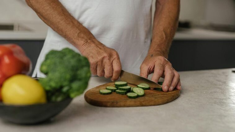 Close-up of adult slicing cucumber on wooden board with fresh vegetables in background.