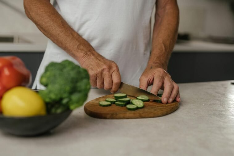 Close-up of adult slicing cucumber on wooden board with fresh vegetables in background.