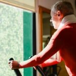 Elderly man working out on exercise bike with towel in a sunny gym.