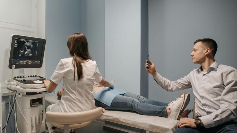 A doctor performs an ultrasound on a pregnant woman, while a man takes a photo in a clinical environment.