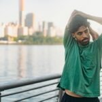 A man in a green shirt stretches outdoors by the waterfront, city skyline in the background.