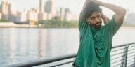 A man in a green shirt stretches outdoors by the waterfront, city skyline in the background.