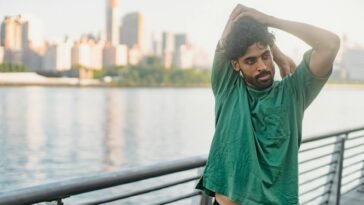A man in a green shirt stretches outdoors by the waterfront, city skyline in the background.
