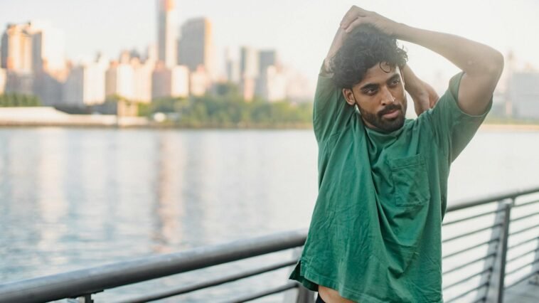 A man in a green shirt stretches outdoors by the waterfront, city skyline in the background.