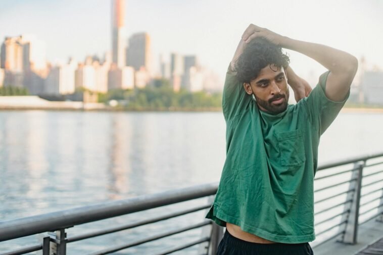 A man in a green shirt stretches outdoors by the waterfront, city skyline in the background.