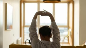 Back view of a man stretching inside a room with a sunny window backdrop.