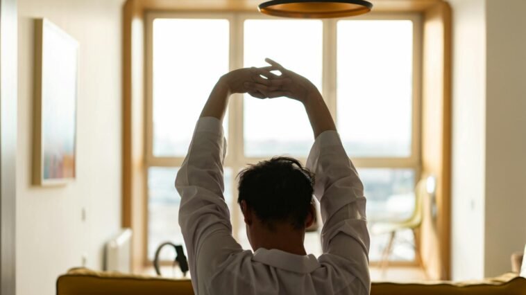 Back view of a man stretching inside a room with a sunny window backdrop.