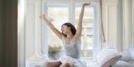 A woman in a gray tank top stretches joyfully on her bed in a bright room, embracing a new day.
