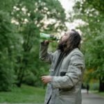 Man with long hair drinks from a green bottle in a lush green park setting.