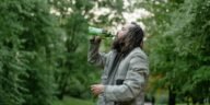 Man with long hair drinks from a green bottle in a lush green park setting.