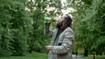 Man with long hair drinks from a green bottle in a lush green park setting.