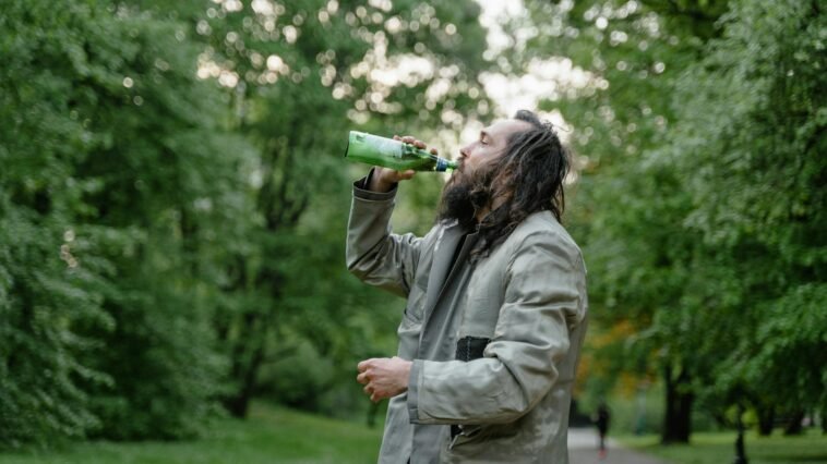 Man with long hair drinks from a green bottle in a lush green park setting.