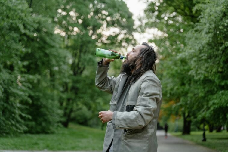 Man with long hair drinks from a green bottle in a lush green park setting.