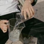 Close-up of a person pouring water from a glass into a pitcher. Outdoors setting.