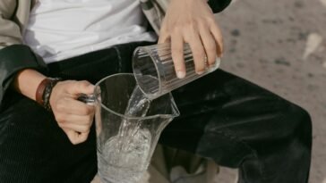 Close-up of a person pouring water from a glass into a pitcher. Outdoors setting.