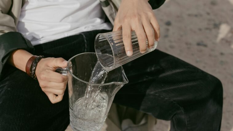 Close-up of a person pouring water from a glass into a pitcher. Outdoors setting.