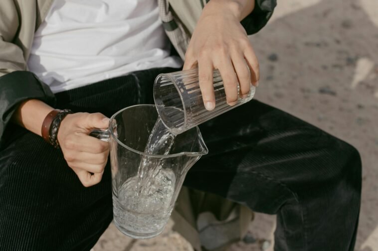 Close-up of a person pouring water from a glass into a pitcher. Outdoors setting.