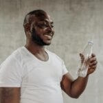 A happy man smiles while holding a bottle of water indoors, embracing healthy living.