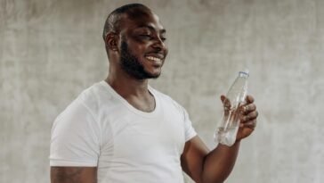 A happy man smiles while holding a bottle of water indoors, embracing healthy living.