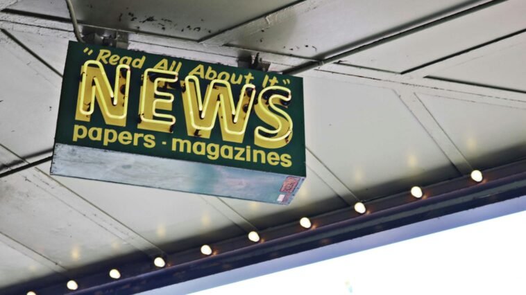 From below of illuminated signboard with news papers magazines inscriptions hanging on metal ceiling on street