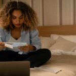 Woman sitting on bed reading documents with laptop, highlighting home office setting.