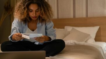 Woman sitting on bed reading documents with laptop, highlighting home office setting.