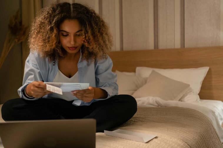 Woman sitting on bed reading documents with laptop, highlighting home office setting.