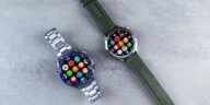 Two smart watches with colorful interfaces displayed on a sleek gray textured surface.