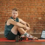 A man sits on an exercise mat with a laptop showing workout routines, suggesting a fitness regimen.