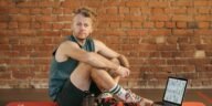 A man sits on an exercise mat with a laptop showing workout routines, suggesting a fitness regimen.