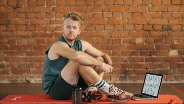 A man sits on an exercise mat with a laptop showing workout routines, suggesting a fitness regimen.