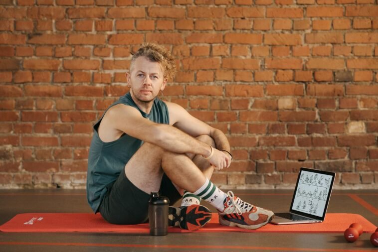 A man sits on an exercise mat with a laptop showing workout routines, suggesting a fitness regimen.