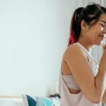 Side view of young Asian female in white shirt standing with closed eyes and enjoying aromatic coffee in morning at home
