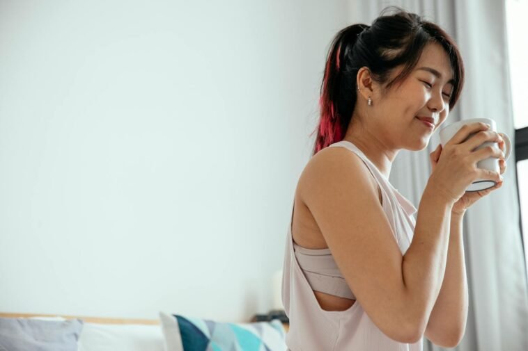 Side view of young Asian female in white shirt standing with closed eyes and enjoying aromatic coffee in morning at home