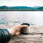 A tranquil lakeside scene with a hand holding a coffee mug on a wooden dock.