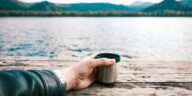 A tranquil lakeside scene with a hand holding a coffee mug on a wooden dock.