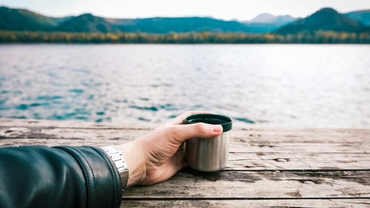 A tranquil lakeside scene with a hand holding a coffee mug on a wooden dock.