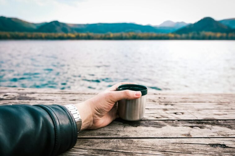 A tranquil lakeside scene with a hand holding a coffee mug on a wooden dock.