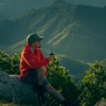 Man enjoys a serene moment in mountain scenery, sipping a drink at sunrise.
