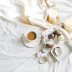 A cozy setting with a cup of coffee, chocolate eggs, and lit candles on a soft white fabric.