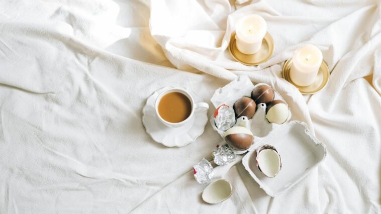 A cozy setting with a cup of coffee, chocolate eggs, and lit candles on a soft white fabric.