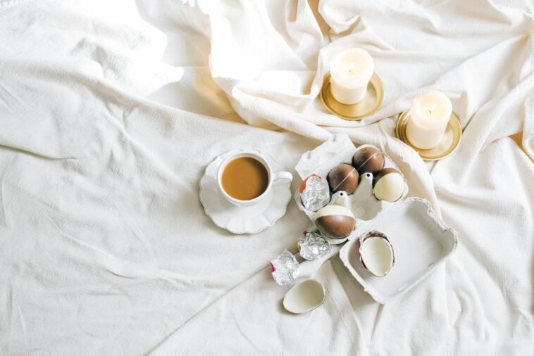 A cozy setting with a cup of coffee, chocolate eggs, and lit candles on a soft white fabric.